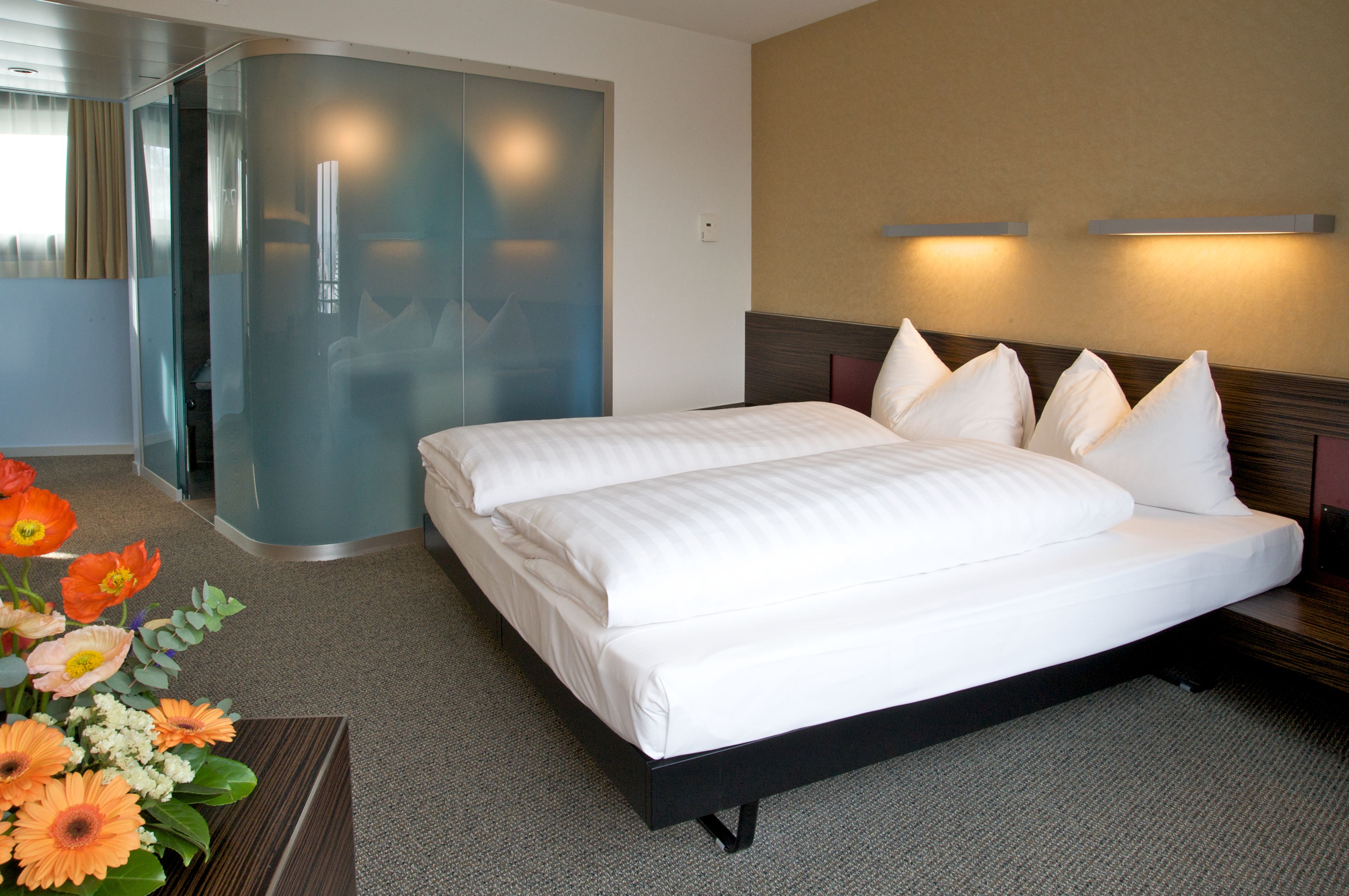 A hotel room with a large bed, a glass partition wall, and a small table with a flower arrangement.