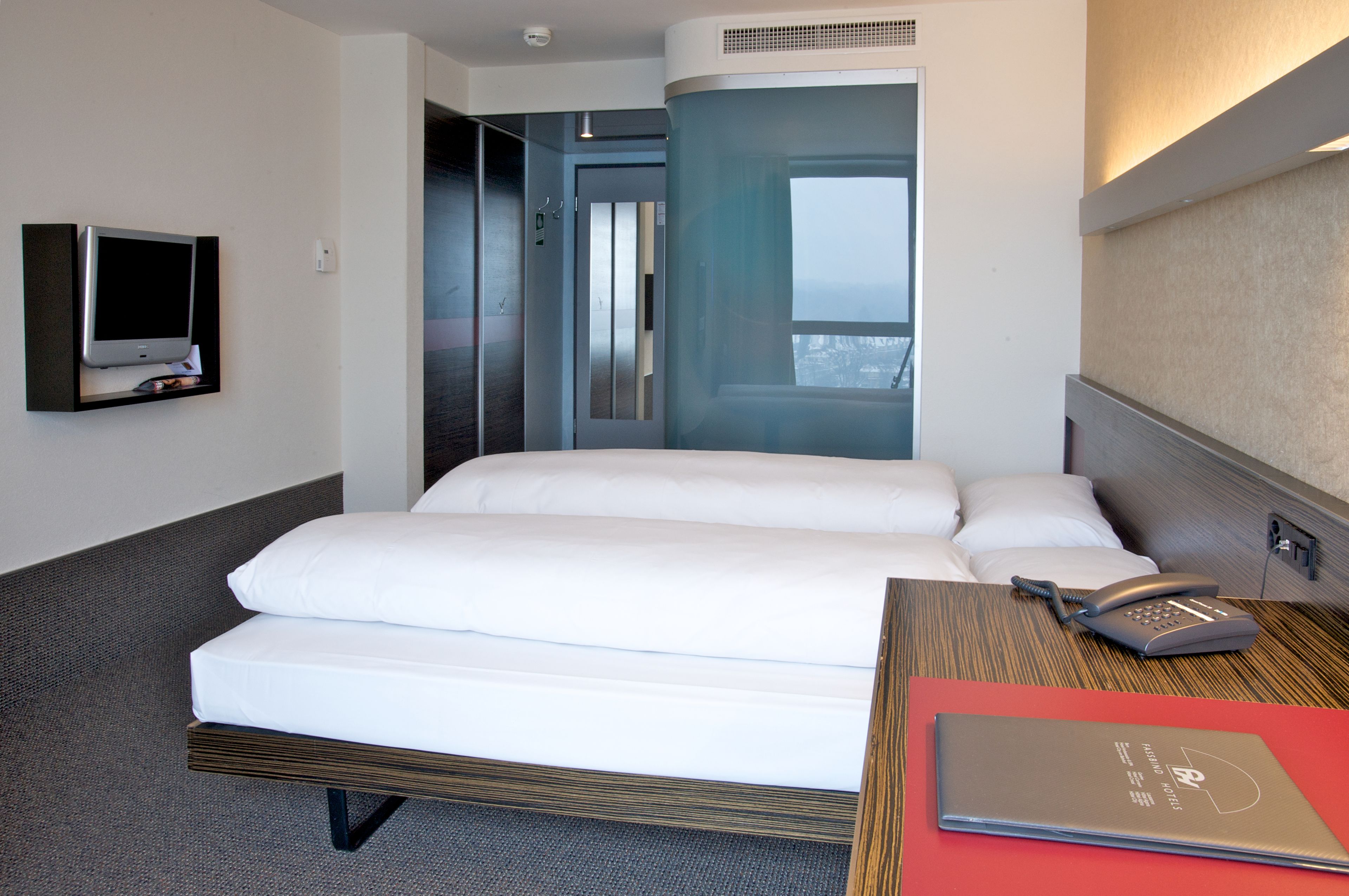 A hotel room with a bed, a bedside table with a phone and a red folder, a TV on the wall, and a view of the city through a window.