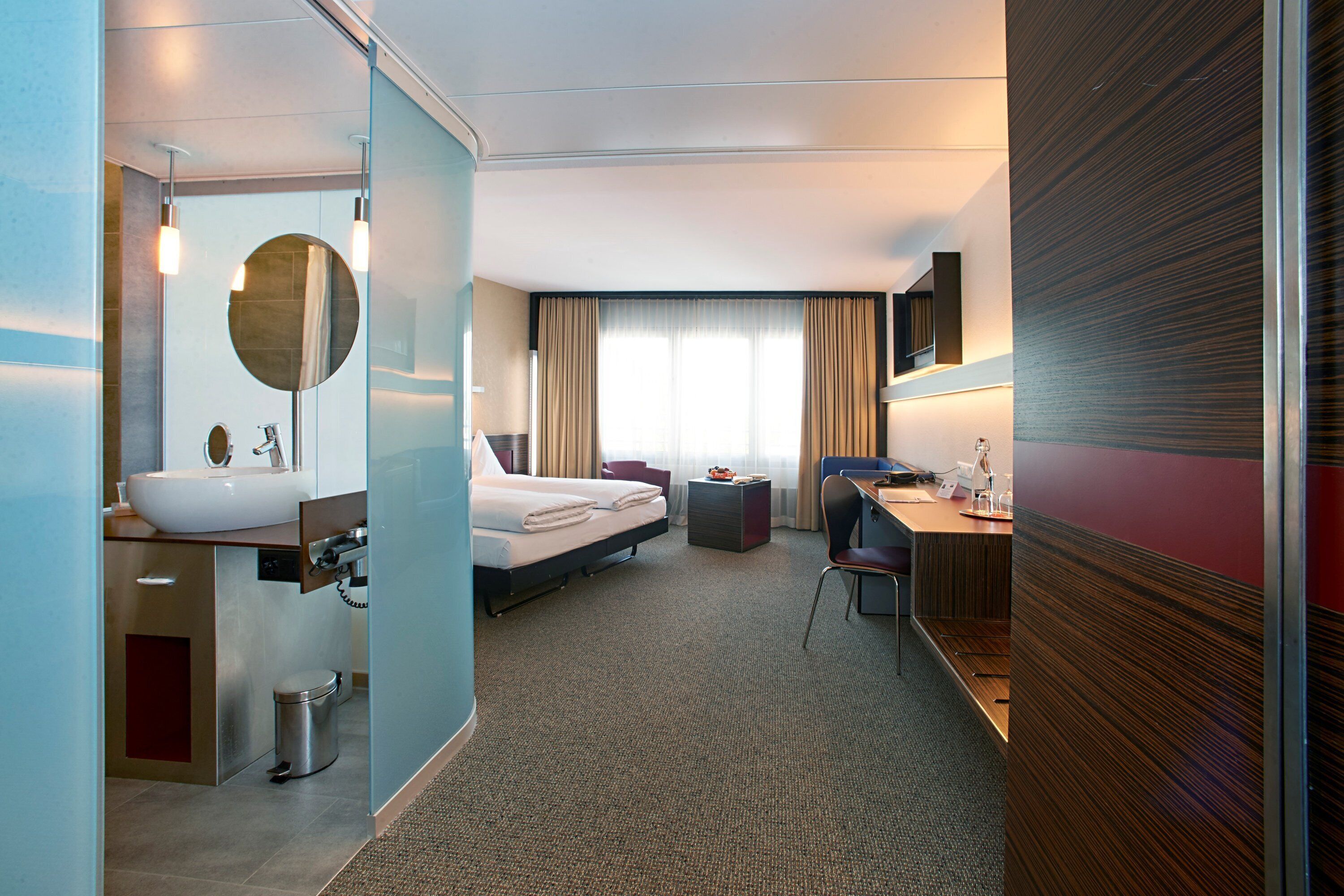 Ein modernes Hotelzimmer mit einem großen Bett, einem Schreibtisch, einem Sessel, einem kleinen Tisch, einem Spiegel und einem Badezimmer mit Waschbecken und Toilette.