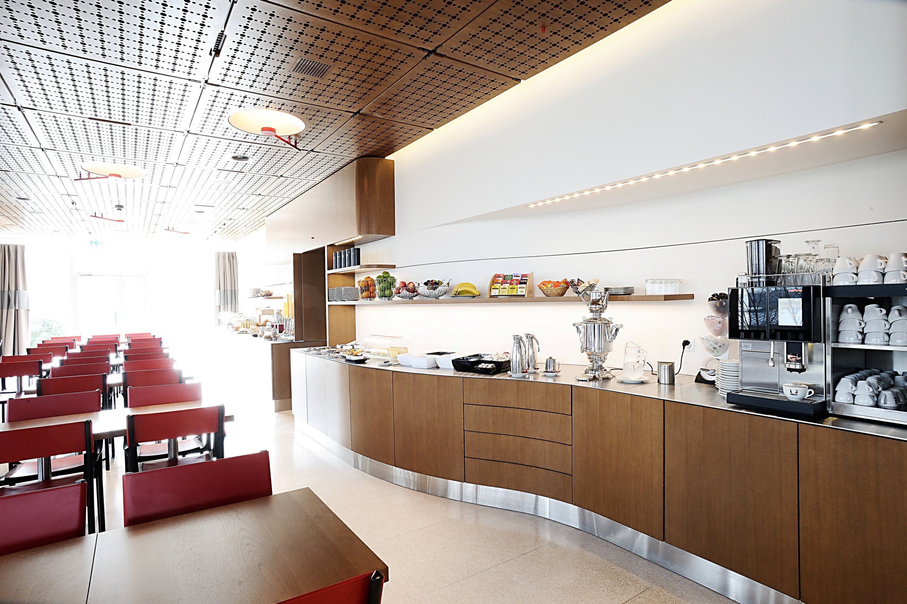 Una cafetería moderna con una barra alargada, armarios de madera y una zona de estar con sillas rojas.