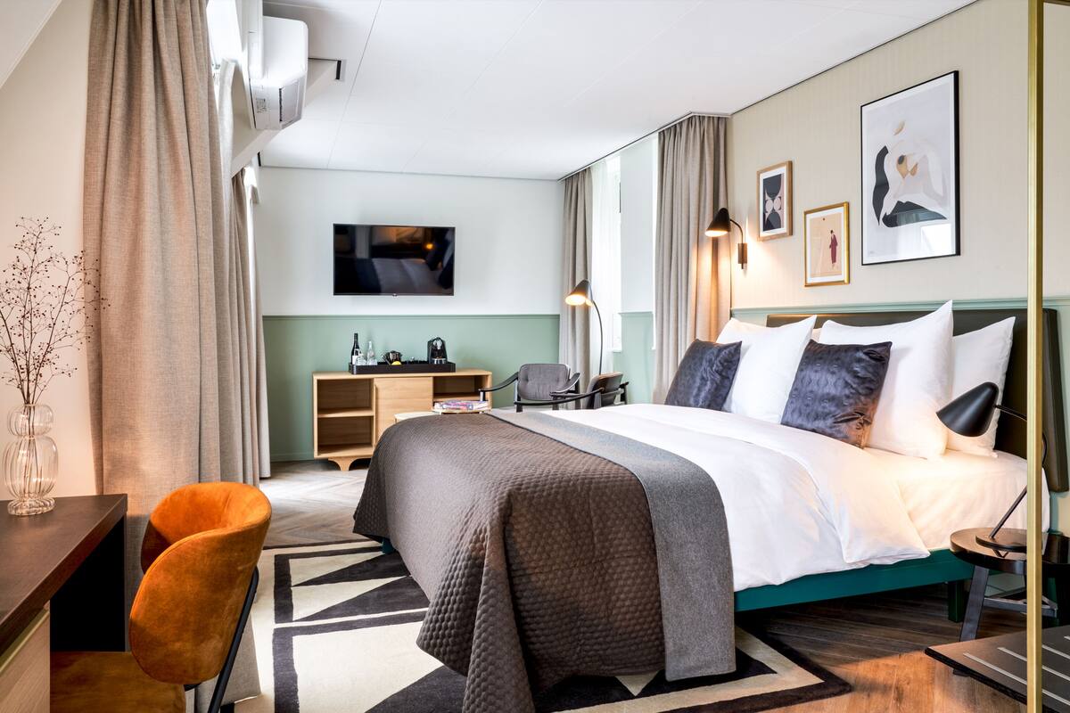 Ein modernes Hotelzimmer mit einem großen Bett, einem Schreibtisch, einem Sessel, einem Fernseher und Kunstwerken an den Wänden.