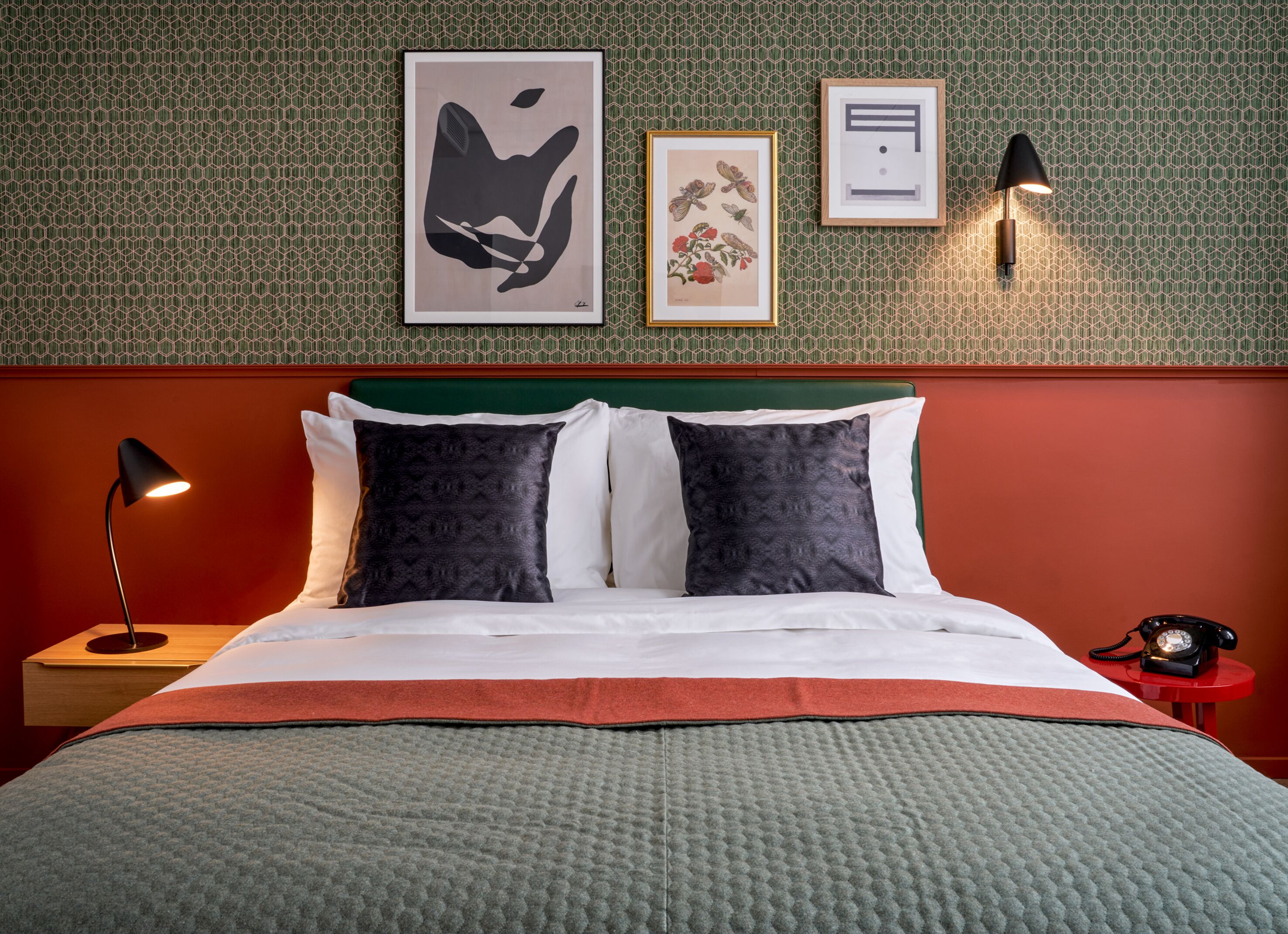 A neatly made bed with a green headboard, two black pillows, and a white pillow. There is a framed artwork above the bed, a wall-mounted lamp on the right, and a vintage telephone on the right side of the bed.