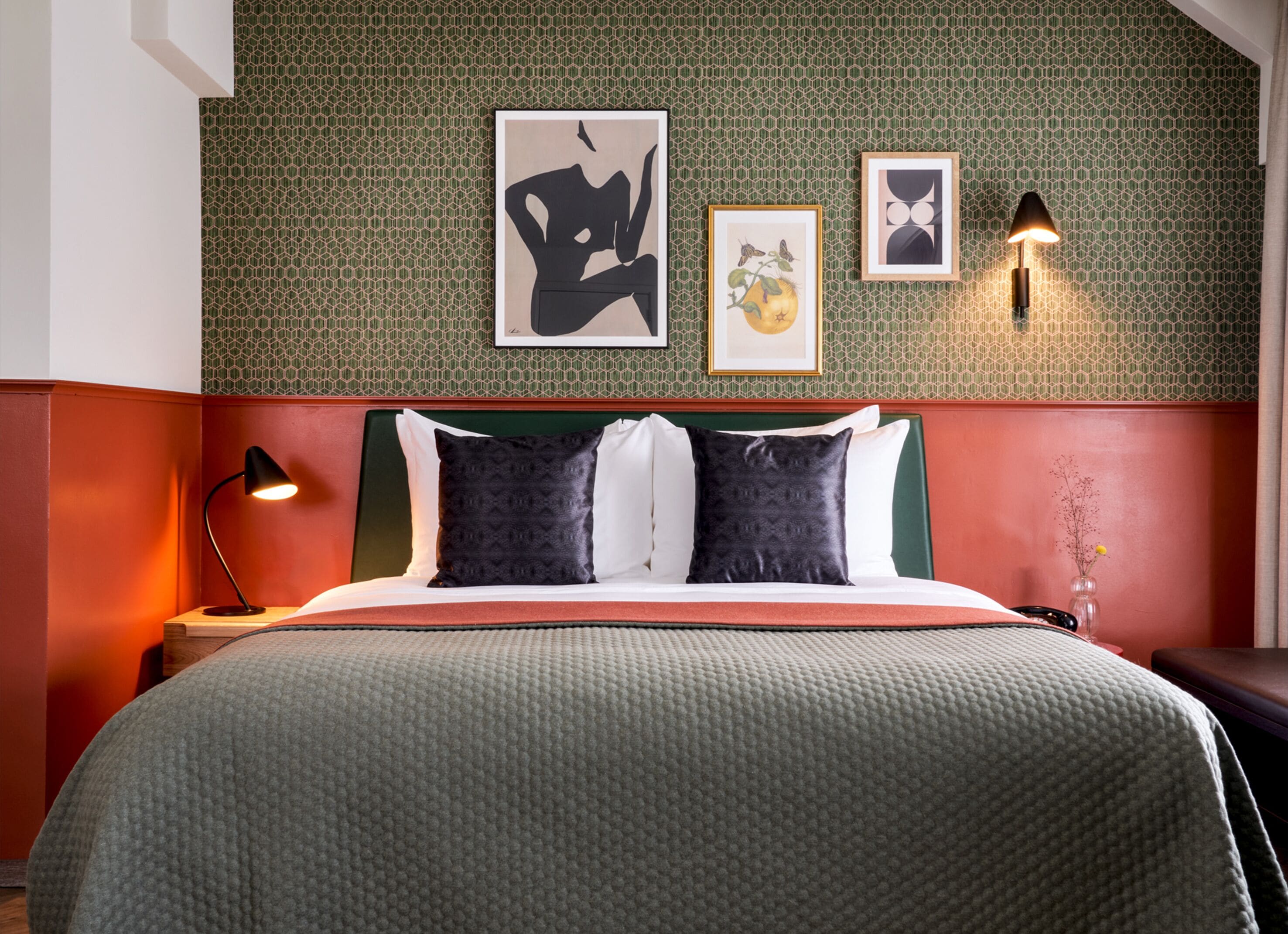 A hotel room with a green textured wall, a red headboard, a bed with a grey quilt, two black pillows, and three framed artworks.