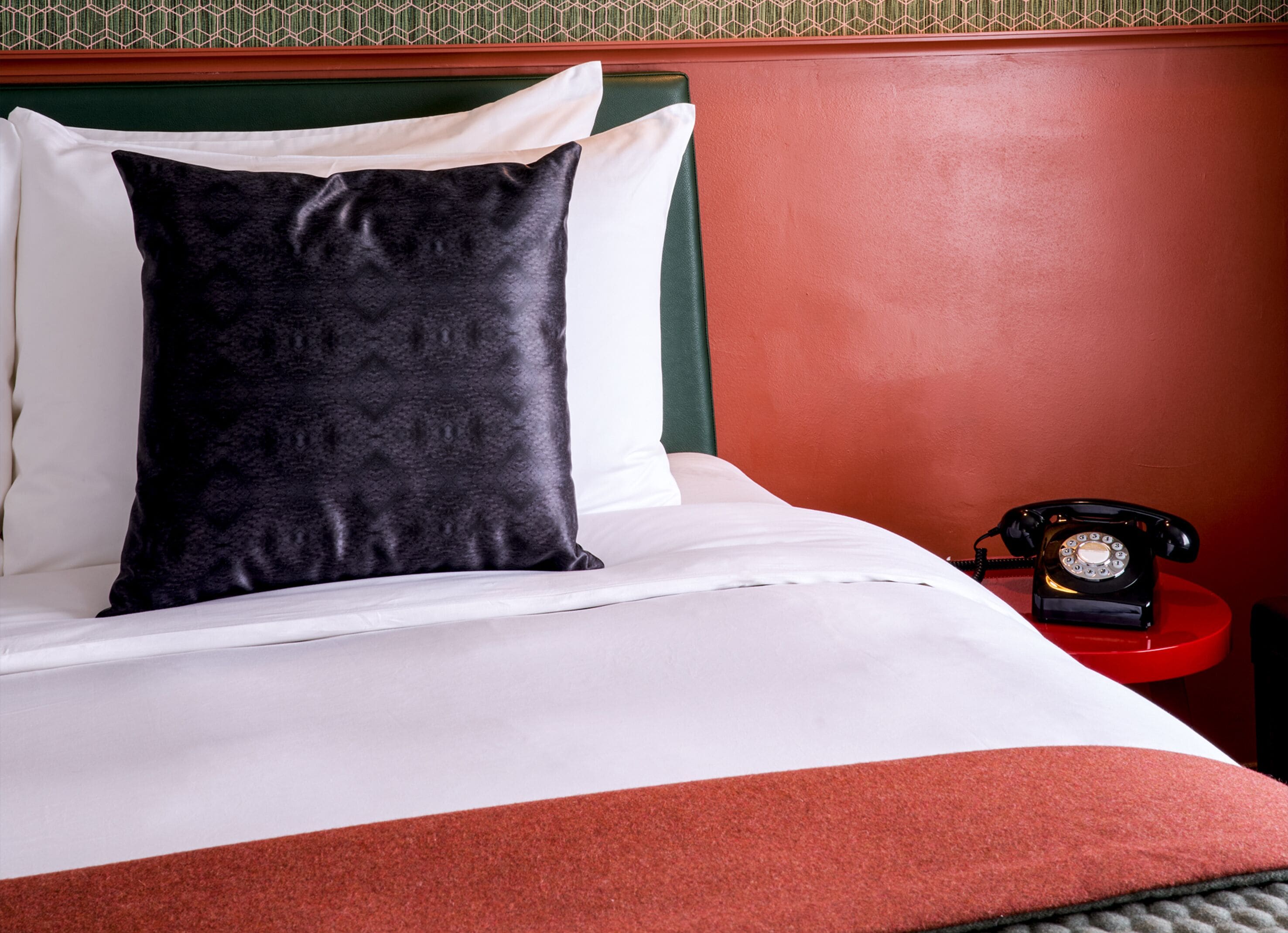 Un lit avec du linge de lit blanc, un oreiller noir et un téléphone rouge posé sur une table de chevet.