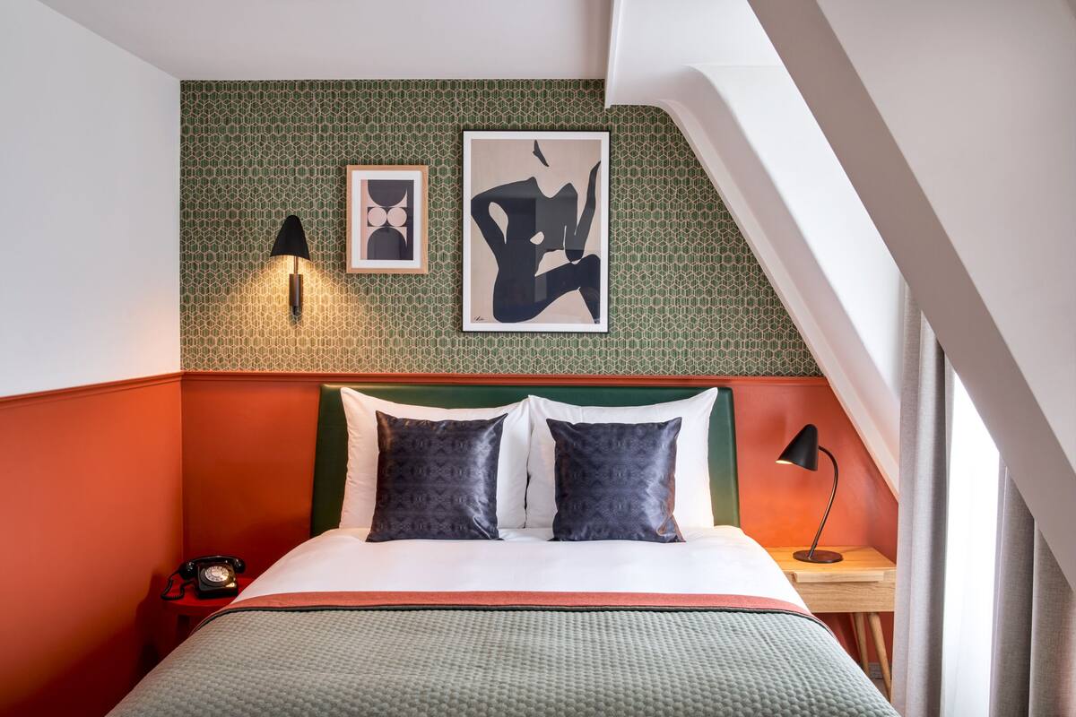 Ein Hotelzimmer mit grüner, strukturierter Wand, einem Bett mit weißen und dunklen Kissen, einem Telefon und einem Nachttisch mit Lampe.