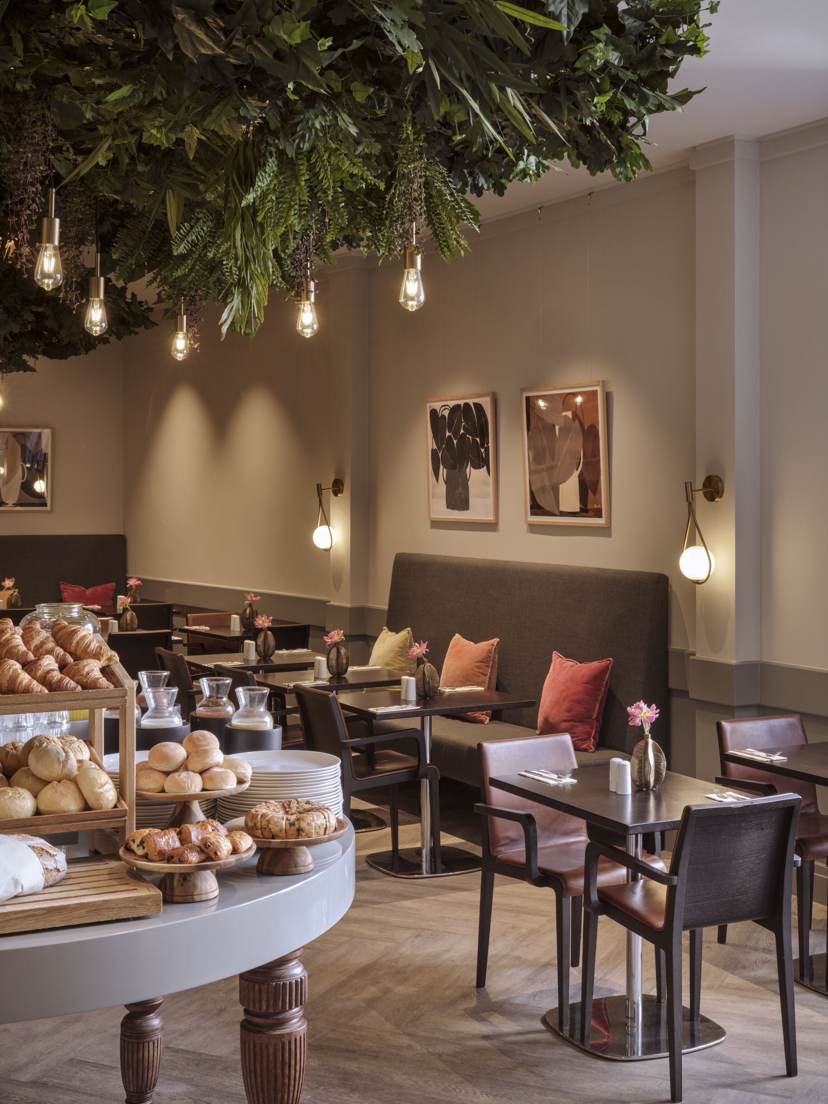 Ein Restaurant mit einer Auswahl an Backwaren auf einem Holzständer, Tische mit Stühlen und hängenden Pflanzen, ergänzt durch Leuchten.
