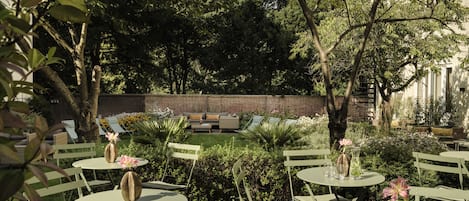 An outdoor seating area with round tables and chairs, surrounded by lush greenery and a brick wall.