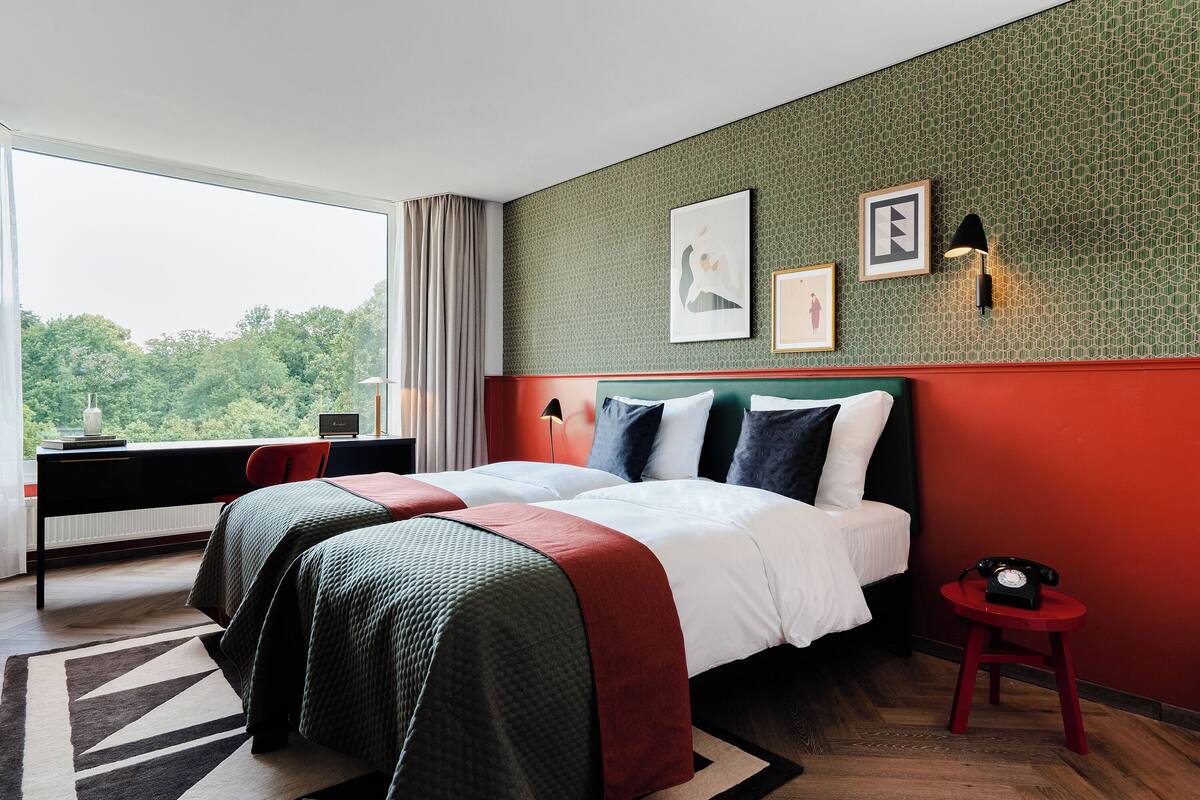 Ein Hotelzimmer mit einem großen Bett, einem Schreibtisch mit Stuhl, einem kleinen Tisch mit Telefon und Blick auf Bäume durch ein Fenster.