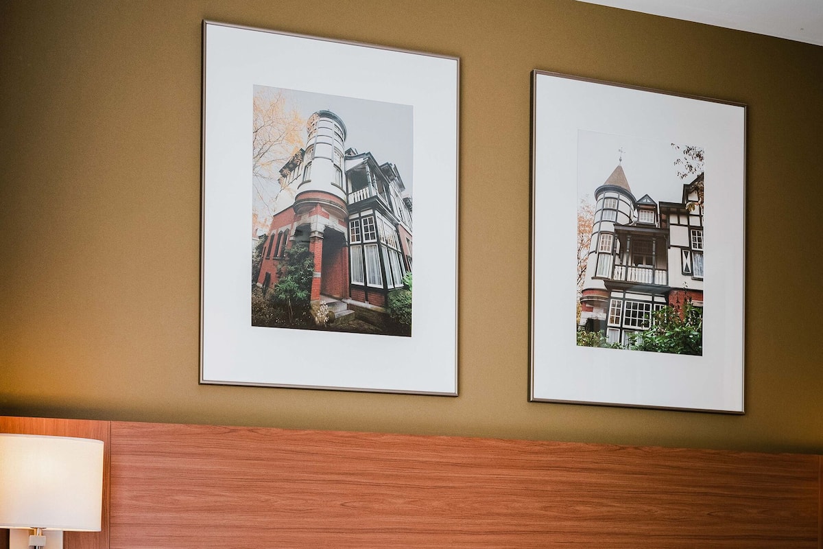 Ein Hotelzimmer mit einem hölzernen Kopfteil, zwei gerahmten Bildern an der Wand, einem Nachttisch mit Lampe und einem Bett mit weißer Bettwäsche und gemusterten Kissen.