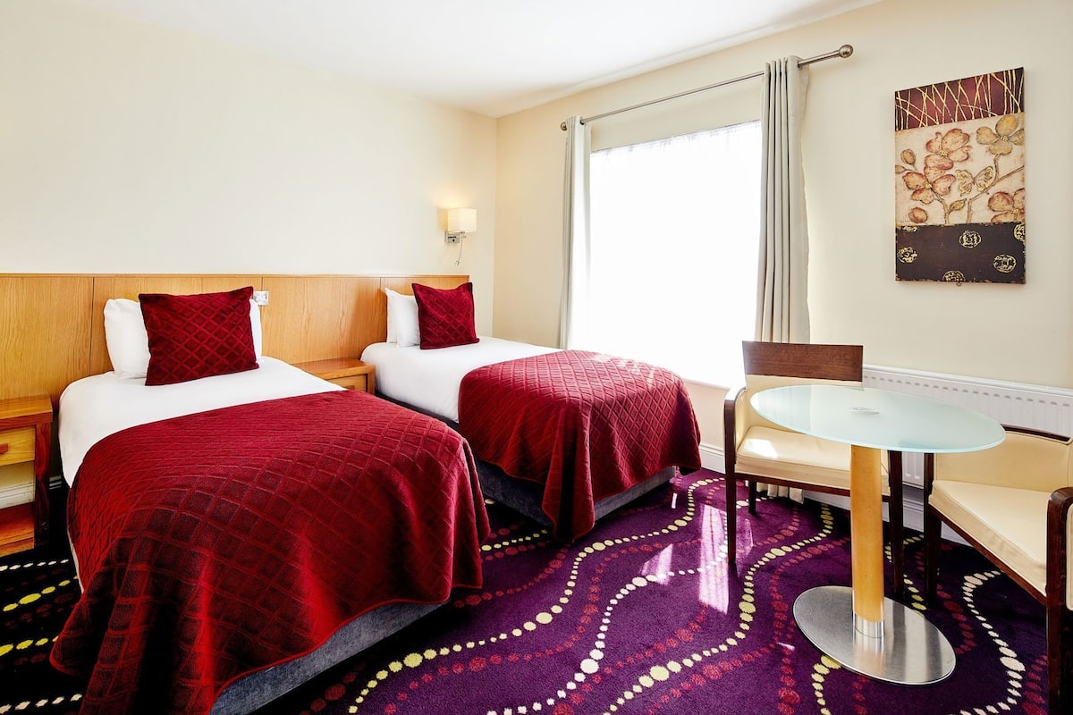 A hotel room with two beds, a round table, a chair, a window with curtains, and a wall-mounted artwork.