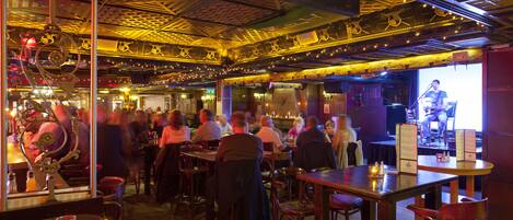 Un bar concurrido con mesas y sillas de madera, un escenario con un artista y un techo con diseño.