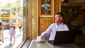 A man sitting at a table with a laptop and a cup of coffee, with a sign advertising Irish coffee in the background. - Arlington Hotel O'Connell Bridge (Dublin)