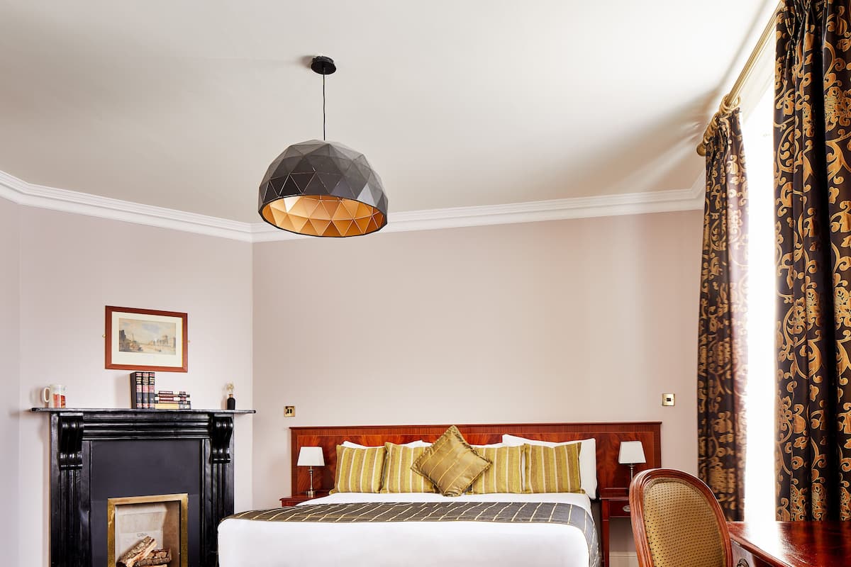 A hotel room with a large bed, a wooden nightstand, a desk with a chair, a hanging light fixture, and a framed picture on the wall.