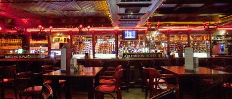 A bar area with multiple tables and chairs, a television screen, and a variety of bottles and glasses on shelves.