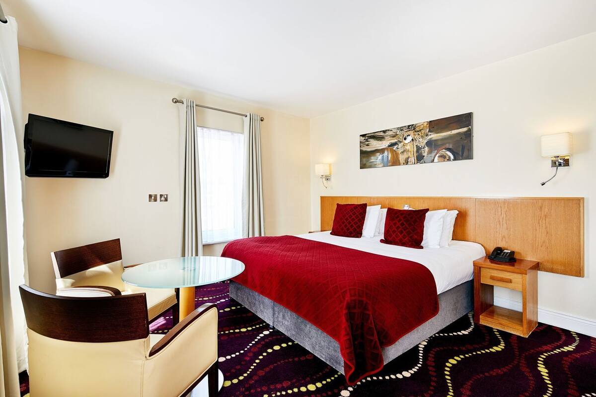 A hotel room with a bed, a desk, a chair, a TV, a painting, and a telephone.