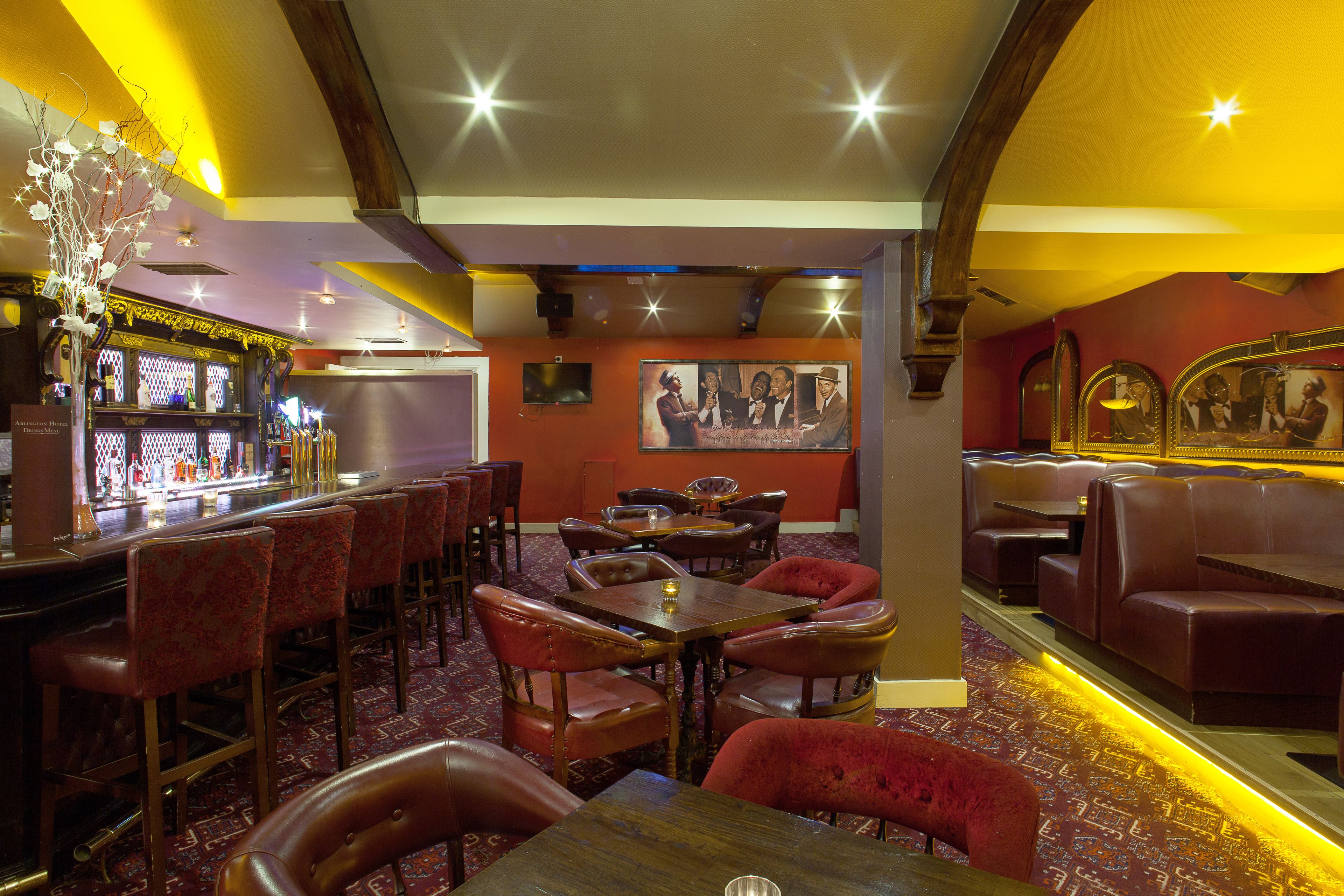 Un restaurant avec des tables en bois et des banquettes rouges, un bar avec des bouteilles exposées, et un mur orné de tableaux.