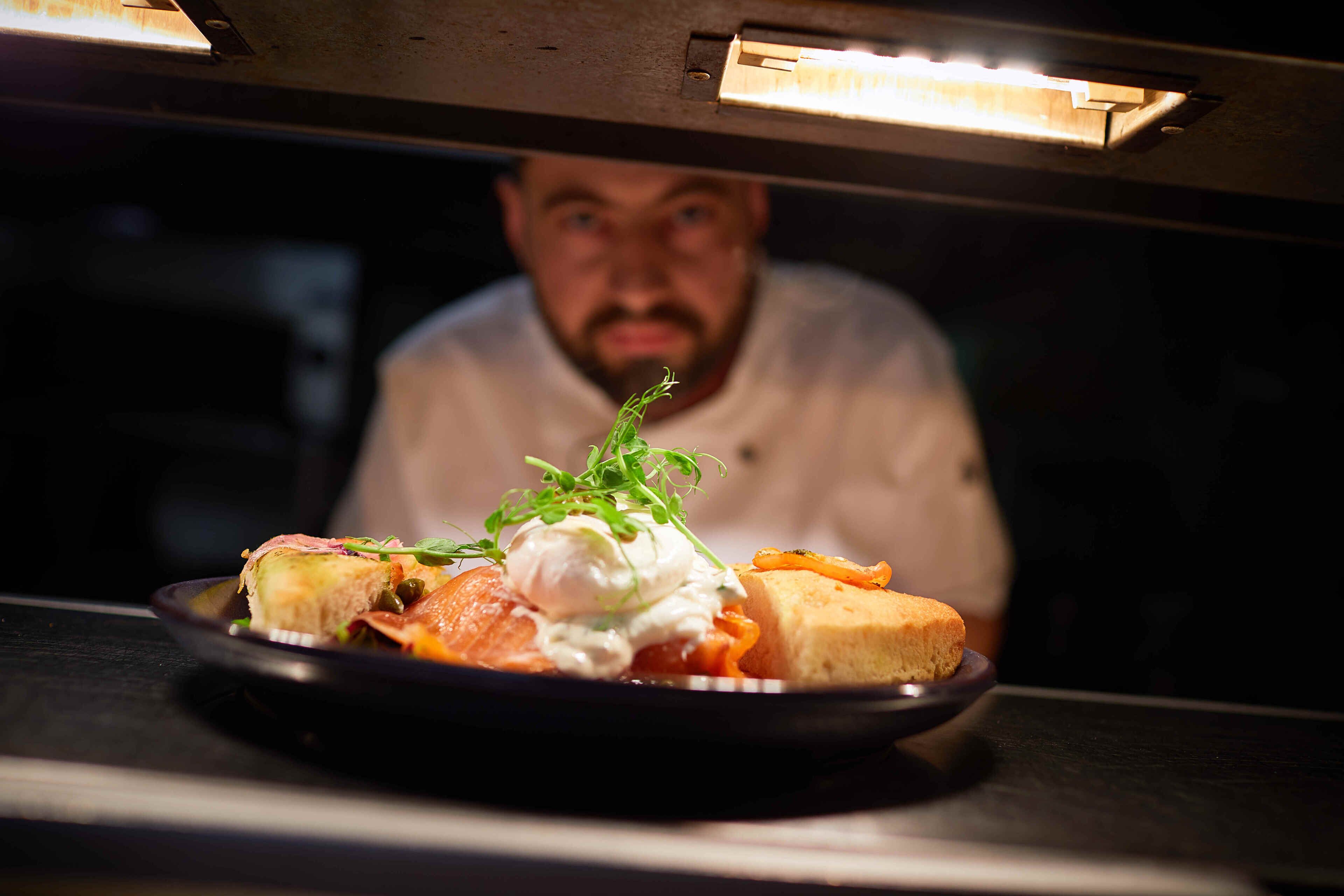 A chef preparing a dish with a plate of food, including a poached egg, smoked salmon, and a slice of bread, in a kitchen setting.