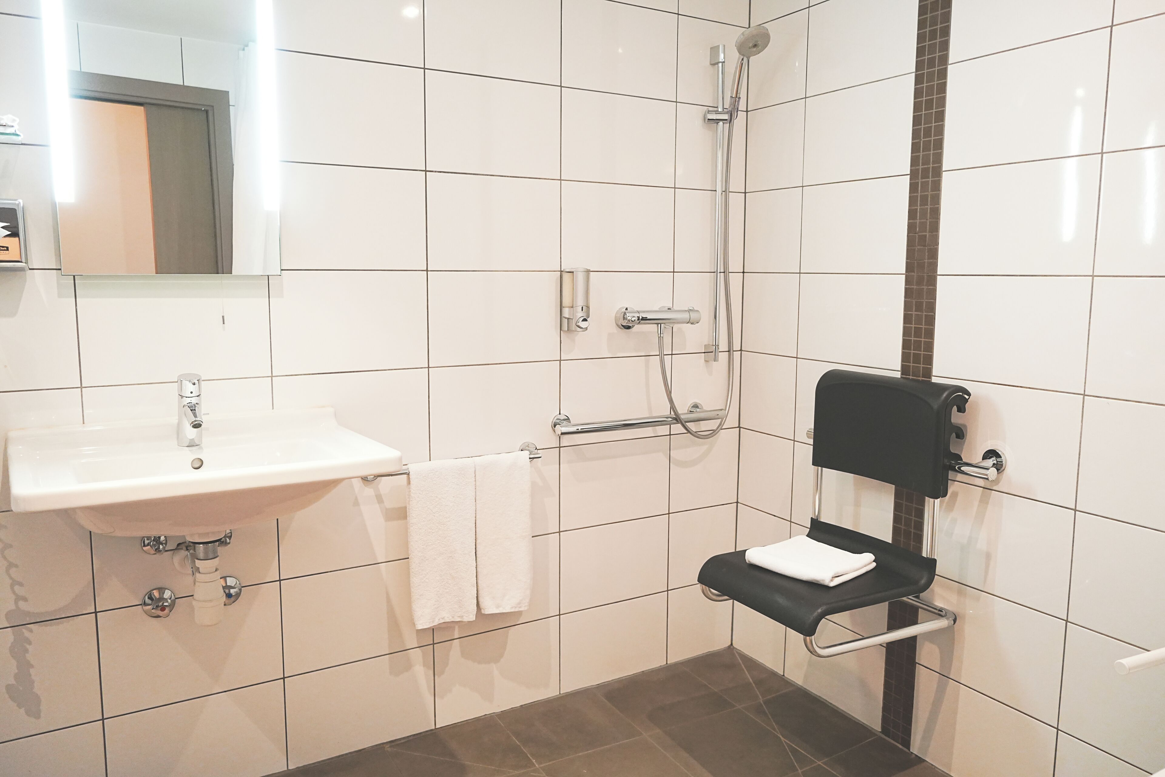 A modern bathroom with a white sink, a mirror, a towel rack, a shower with a handheld showerhead, and a black toilet with a grab bar.
