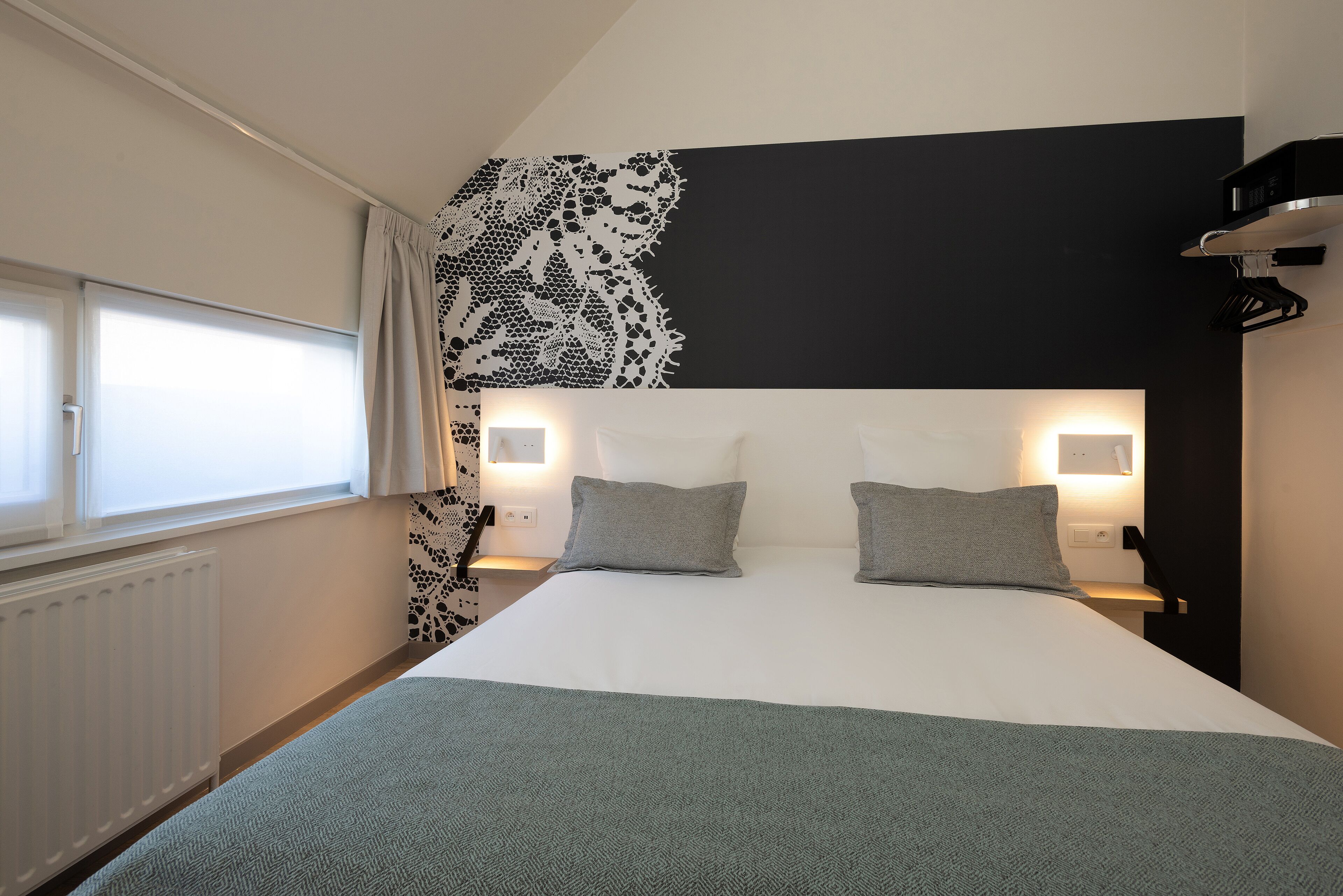 Habitación de hotel con una cama grande, dos mesitas de noche con lámparas, una ventana con cortinas y un papel tapiz blanco y negro.