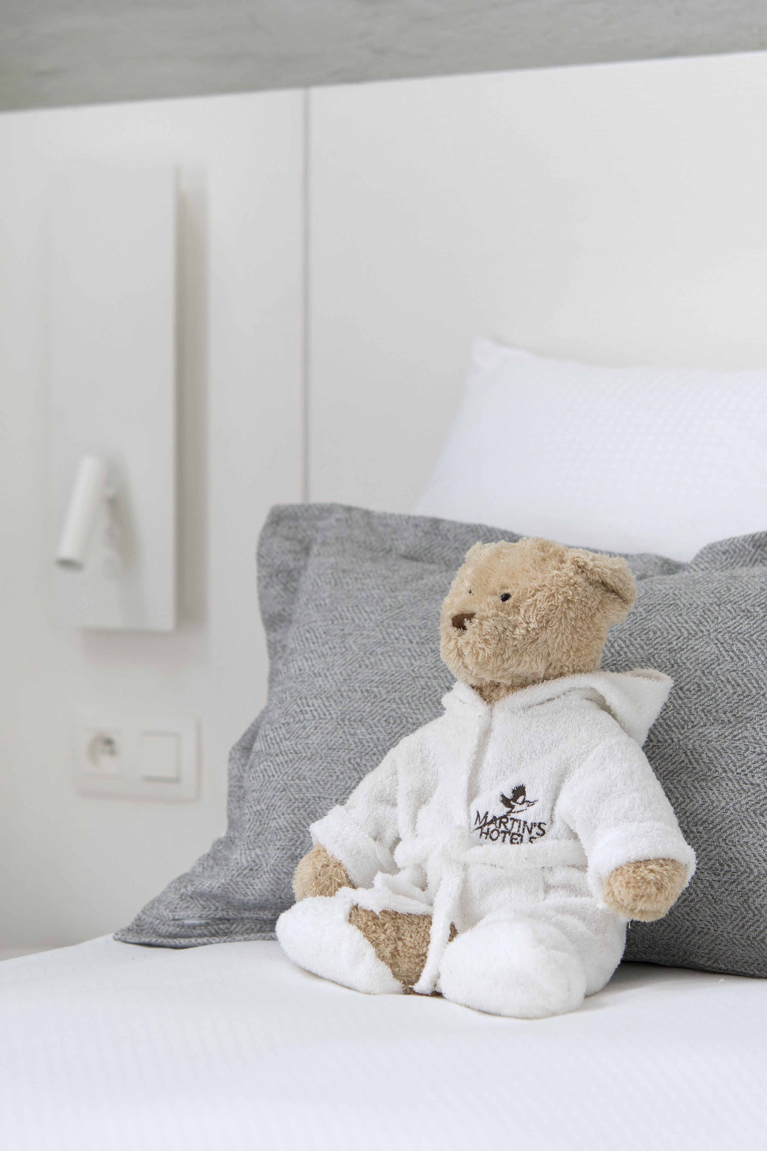 Un osito de peluche con bata blanca sentado en una cama con almohadas grises.