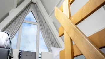 Une pièce avec un plafond en pente, un escalier en bois, un canapé en osier, un fauteuil gris et une fenêtre avec des rideaux.