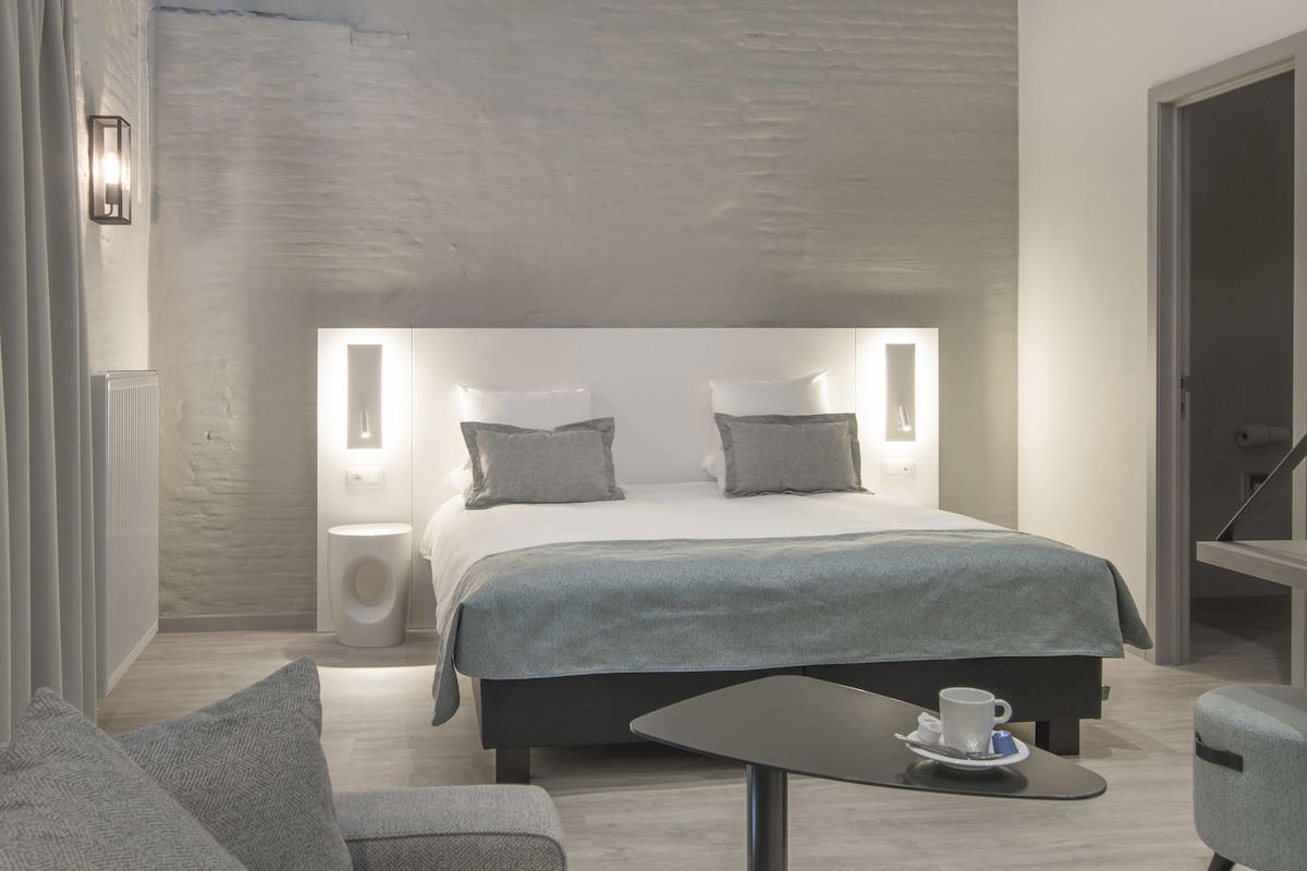 A modern hotel room with a large bed, bedside lamps, a small table with a cup, and a chair.