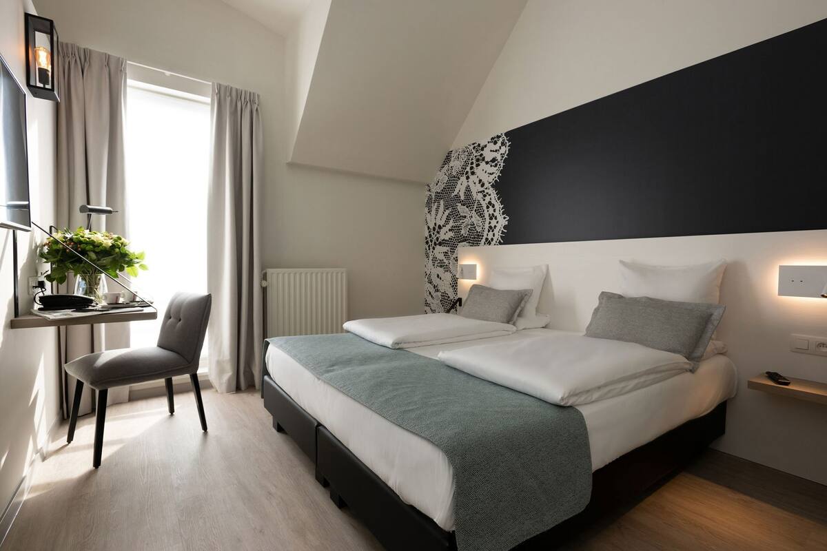 Une chambre moderne comprenant un lit, des tables de chevet, un bureau avec une chaise, un radiateur et une fenêtre avec des rideaux.