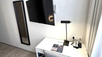 Hotelkamer met een wit bureau, een flatscreen televisie aan de muur, een bureaulamp en een klein prullenbakje.