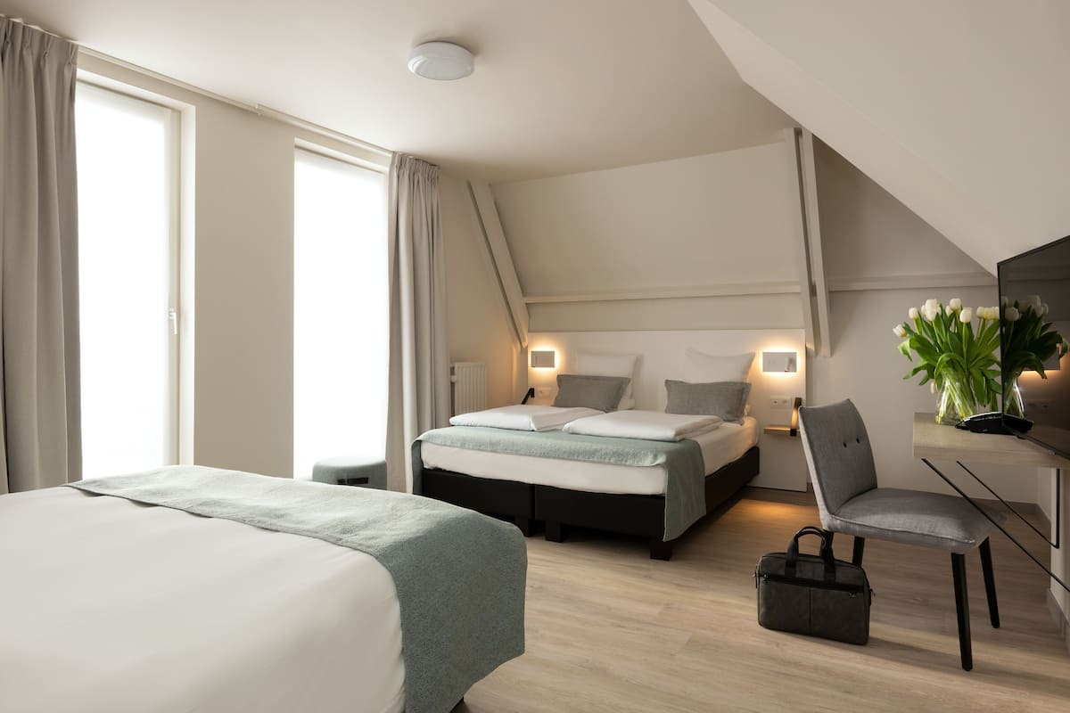 Une chambre d’hôtel moderne avec un grand lit, un bureau et une chaise. On y trouve une fenêtre avec des rideaux, une table de chevet avec une lampe et une plante sur le bureau.