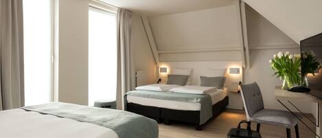 A modern hotel room with a large bed, a desk, and a chair. There is a window with curtains, a bedside table with a lamp, and a plant on the desk.