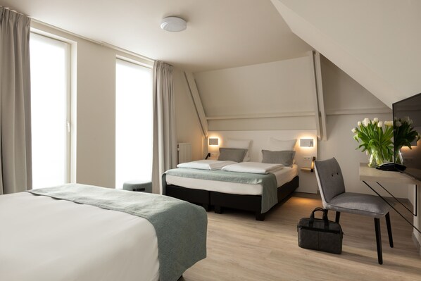 A modern hotel room with a large bed, a desk, and a chair. There is a window with curtains, a bedside table with a lamp, and a plant on the desk. - Martin's Brugge (Bruges)