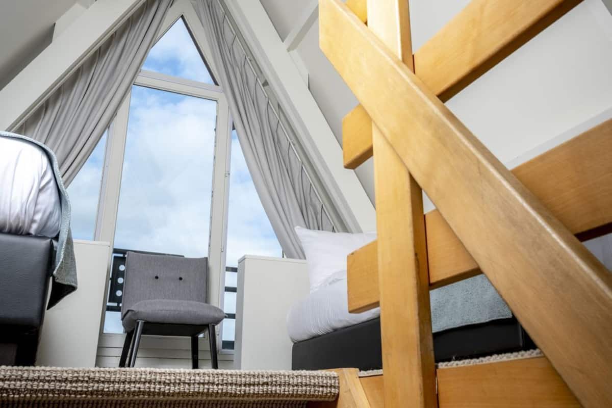 Una habitación con techo alto, una escalera de madera, un ventanal con cortinas, una silla gris y un sofá tejido.