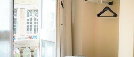 A bedroom with a bed, a window, a hanging coat rack, and a shelf with a small item.