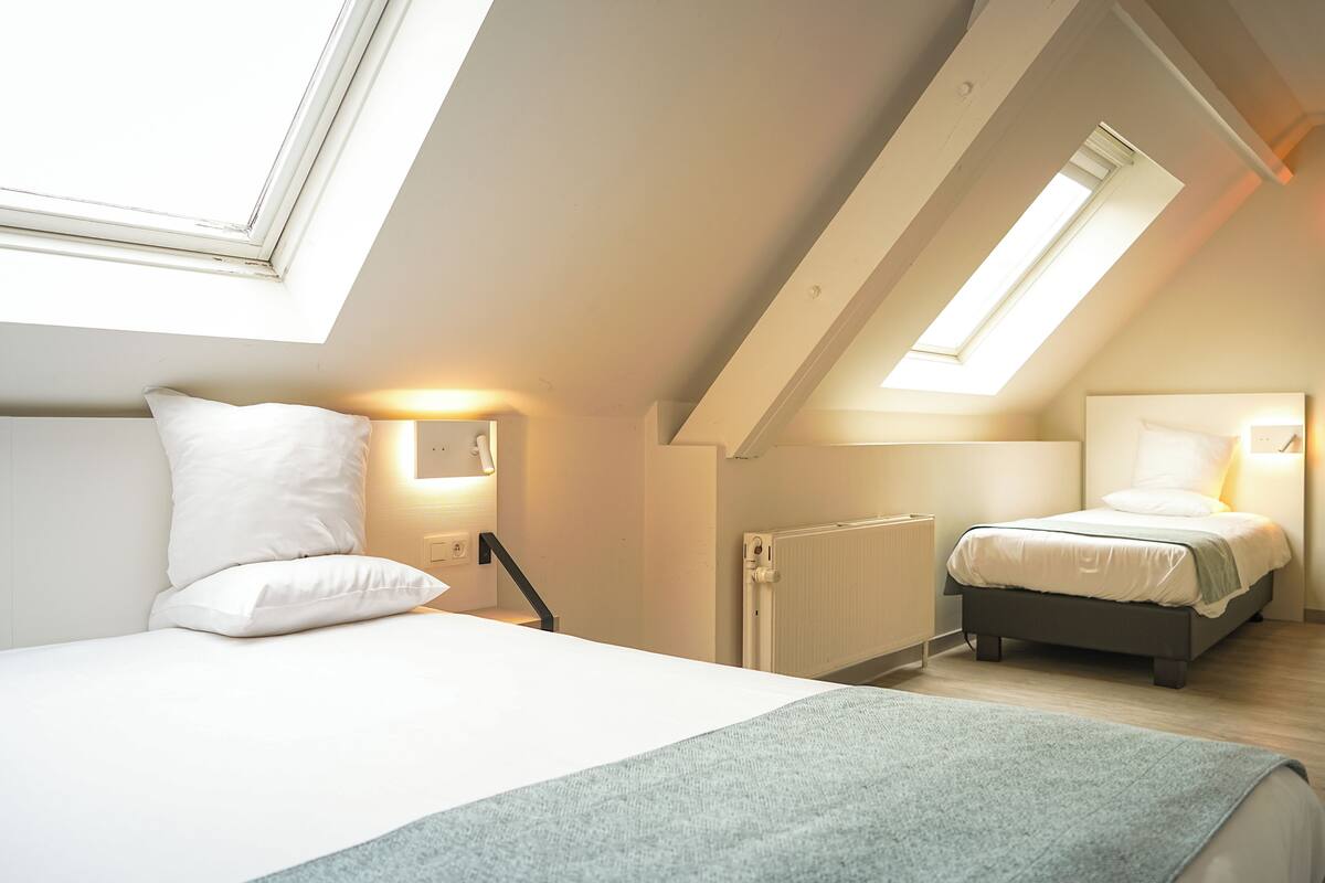 Une chambre avec un plafond en pente, des fenêtres de toit, un lit avec une literie blanche, une table de chevet et une commode.