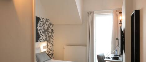 A modern hotel room with a bed, a desk with a chair, a television, and a small stool.