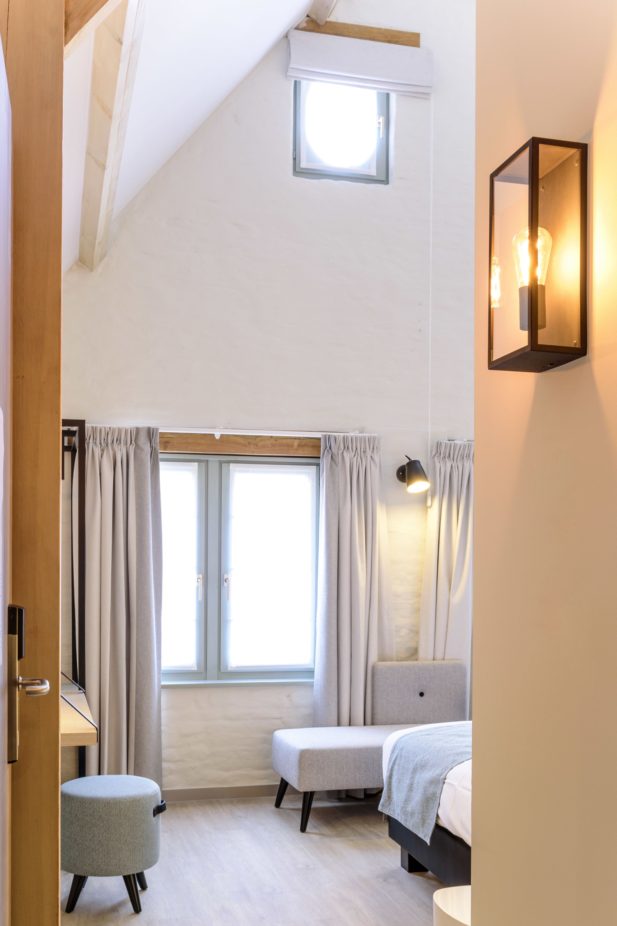 A bedroom with a sloped ceiling, a small skylight, a bed, a bench, a desk, and a wall-mounted light fixture.