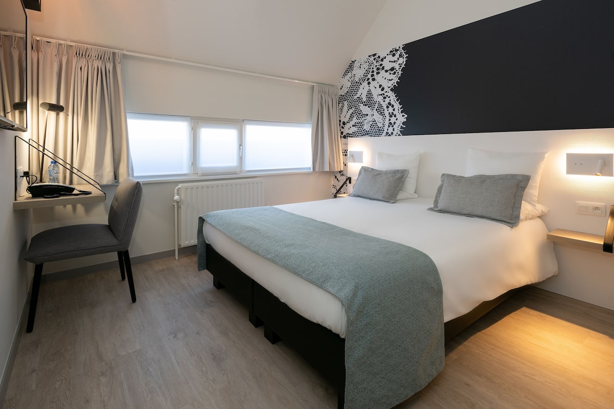 Une chambre d’hôtel moderne, équipée d’un lit, d’une table de chevet, d’une chaise et d’une fenêtre avec des rideaux.