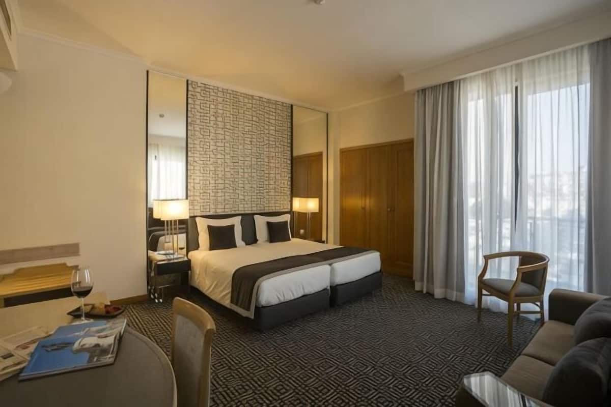 A hotel room with a large bed, a desk with a chair, a dining table, and a window with curtains.