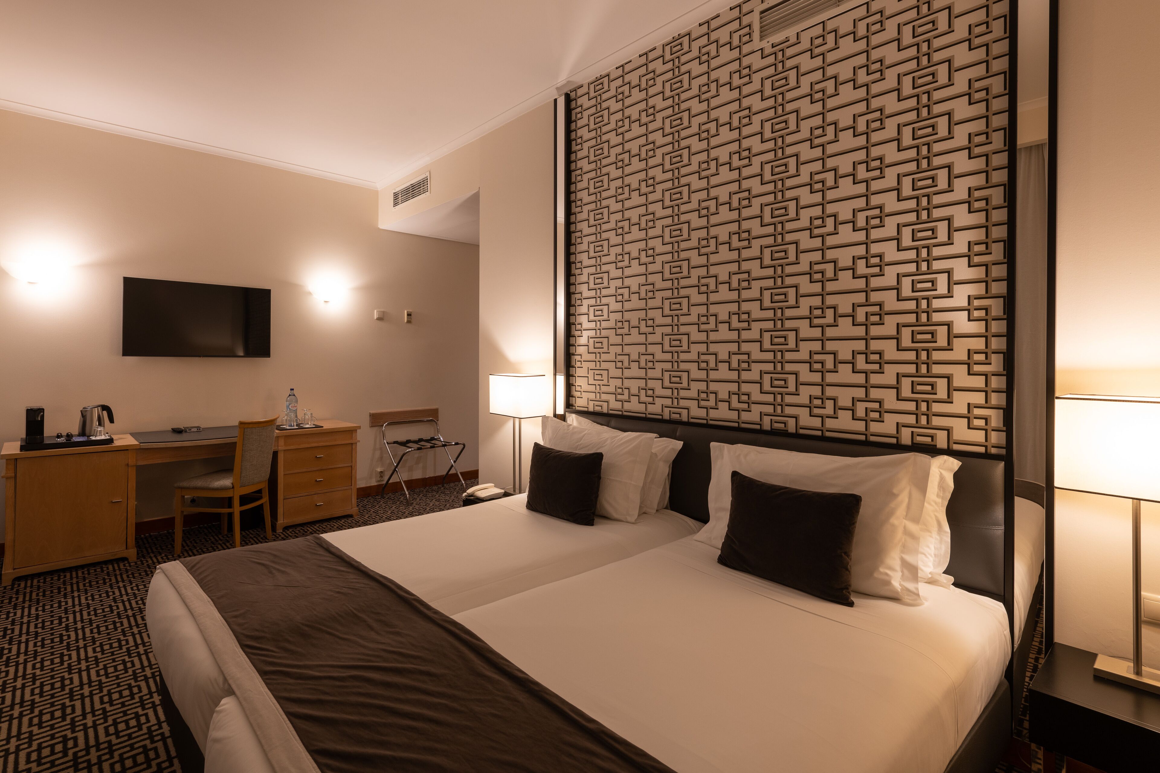 A hotel room with a large bed, a desk with a chair, a TV mounted on the wall, and a patterned headboard.