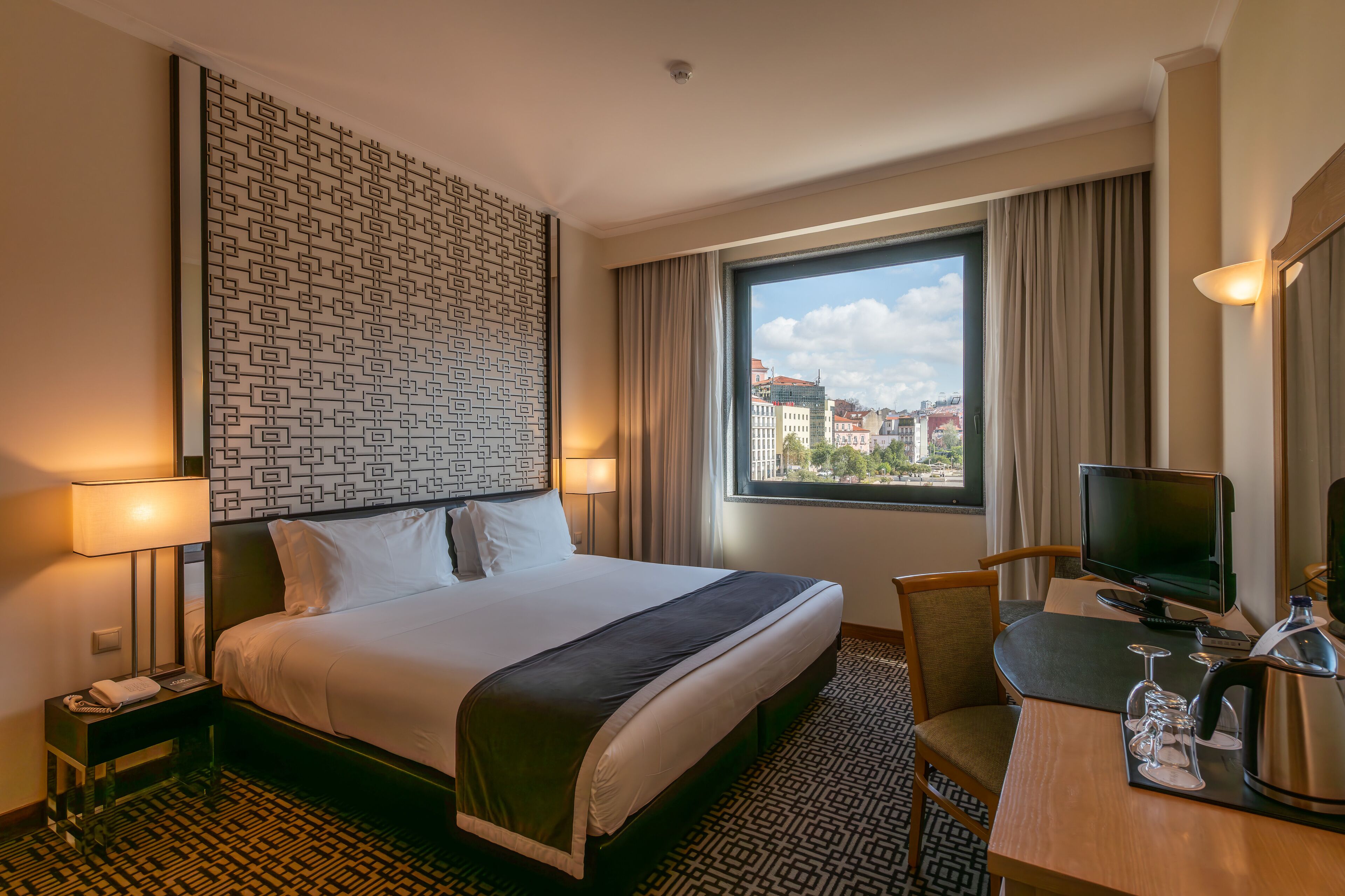 A hotel room with a large bed, a desk with a computer, a chair, a television, and a window with a city view.