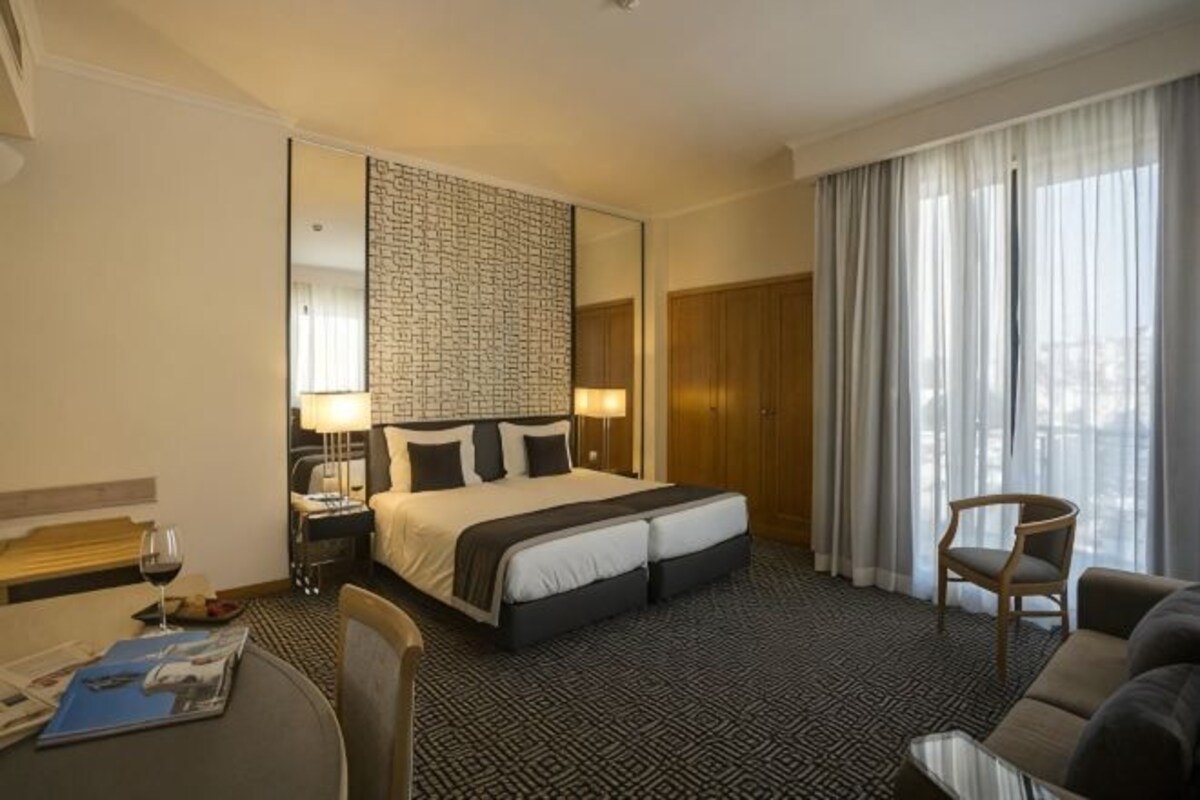 Quarto de hotel com uma cama grande, uma escrivaninha com cadeira, uma mesa de jantar e uma janela com cortinas.
