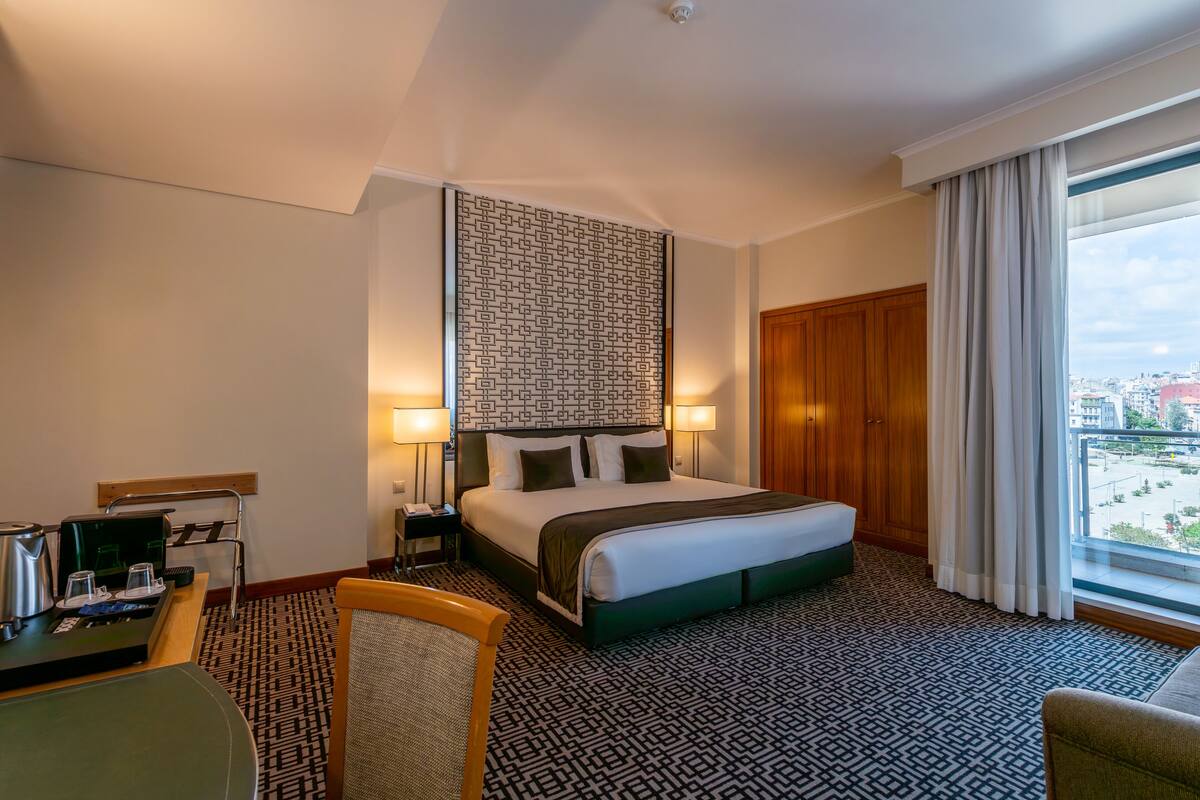 A hotel room with a large bed, two bedside lamps, a desk with a coffee maker, a chair, and a view of the city through a large window.