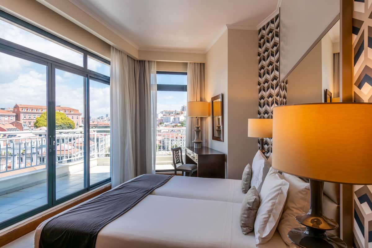A hotel room with a large bed, a desk, and a chair. There is a window with a view of buildings and a cityscape.