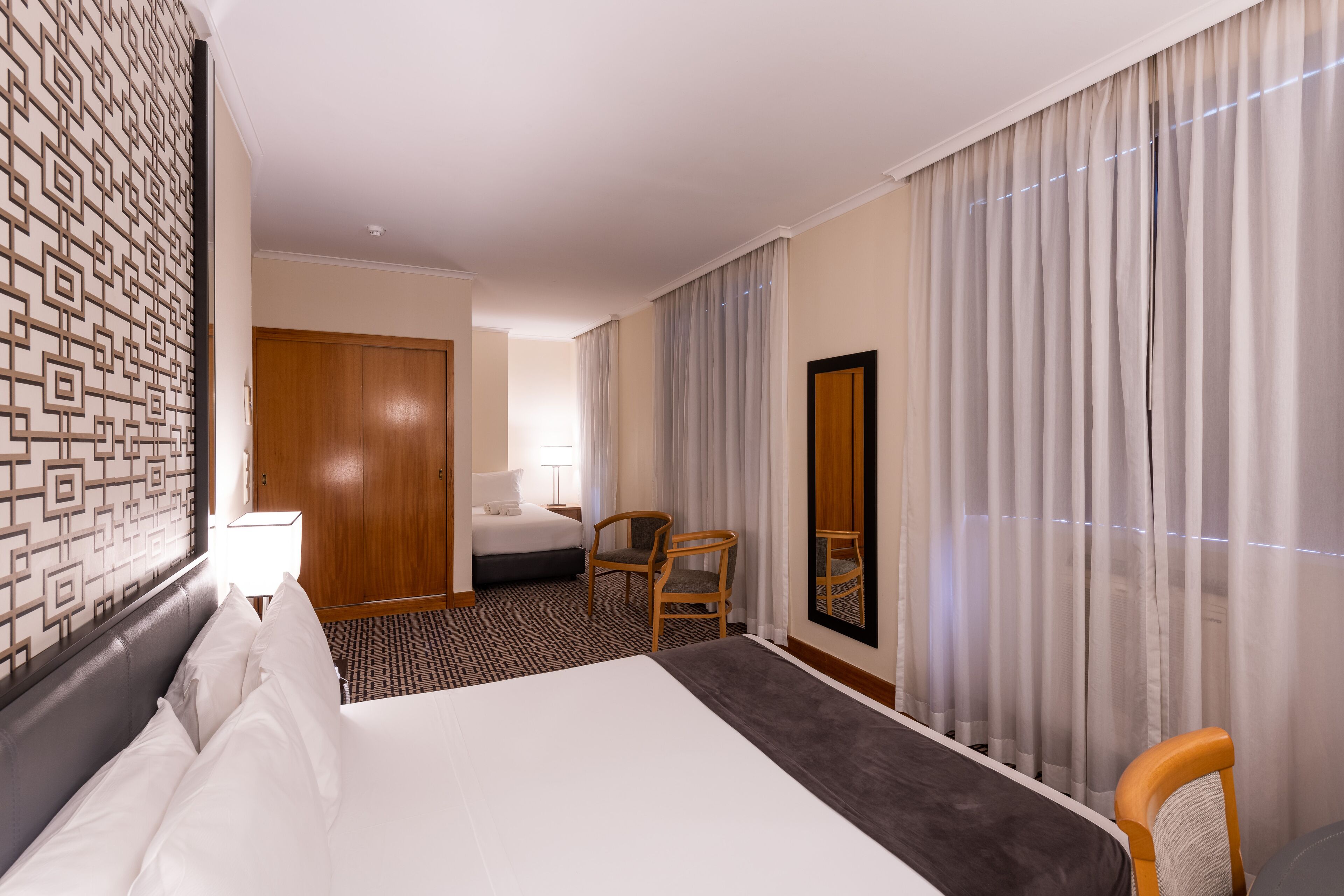 A hotel room with a large bed, a wooden wardrobe, a desk with a chair, and a large window with curtains.
