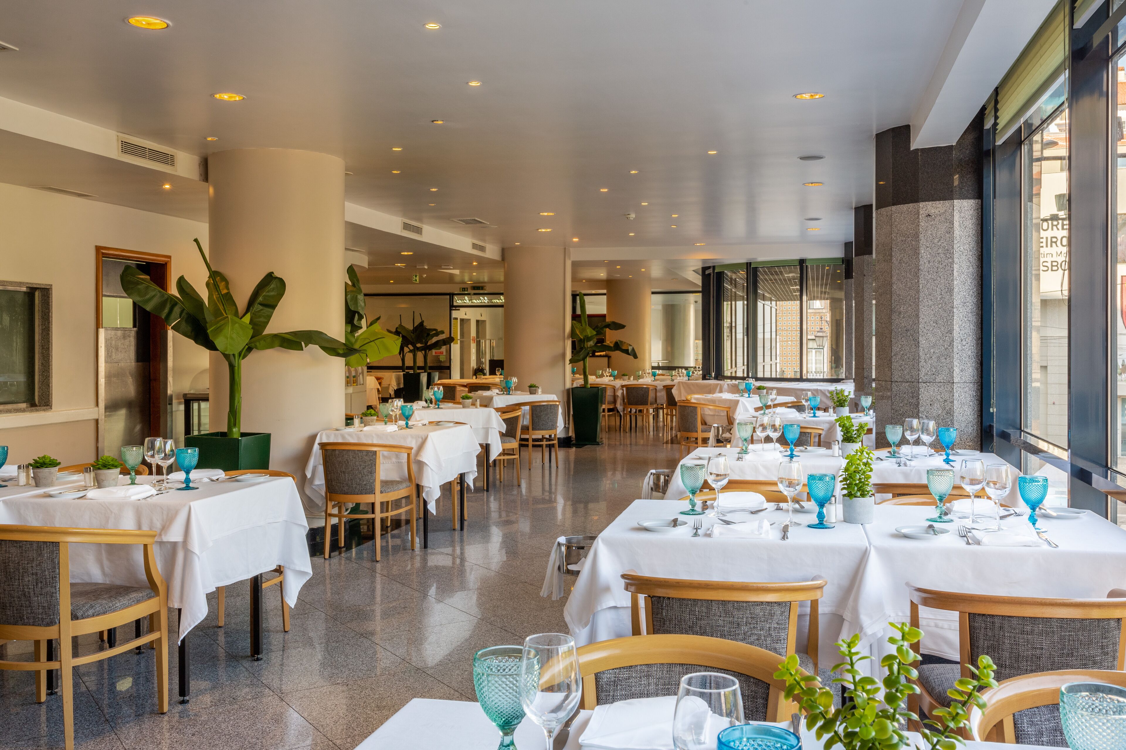 A spacious restaurant with multiple tables set for dining, featuring a modern interior design with large windows and indoor plants.