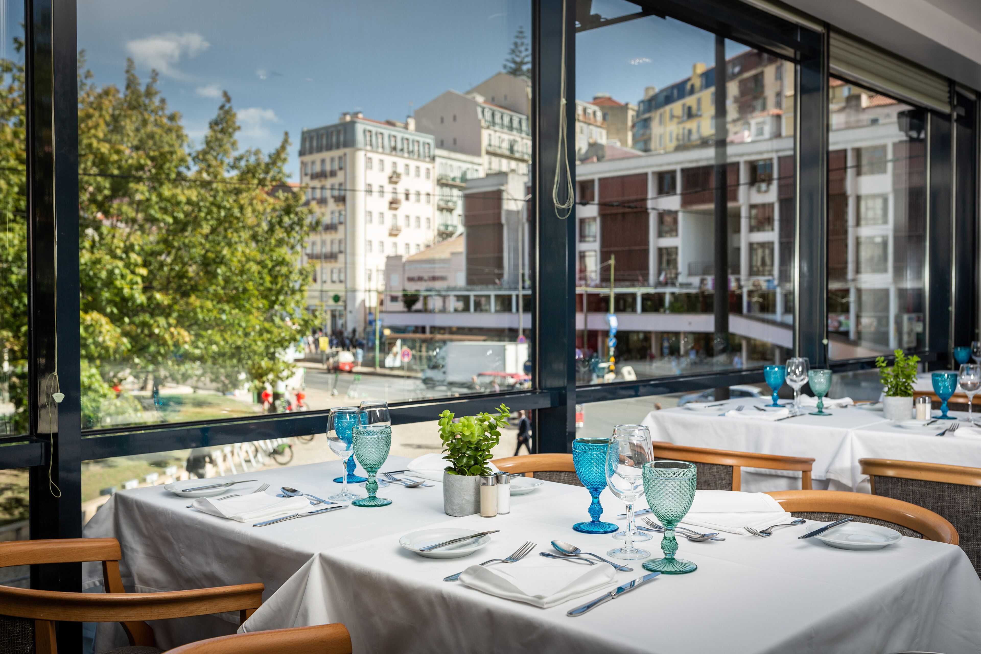 Ein Restaurant mit gedeckten Tischen, große Fenster mit Blick auf Gebäude und Bäume.