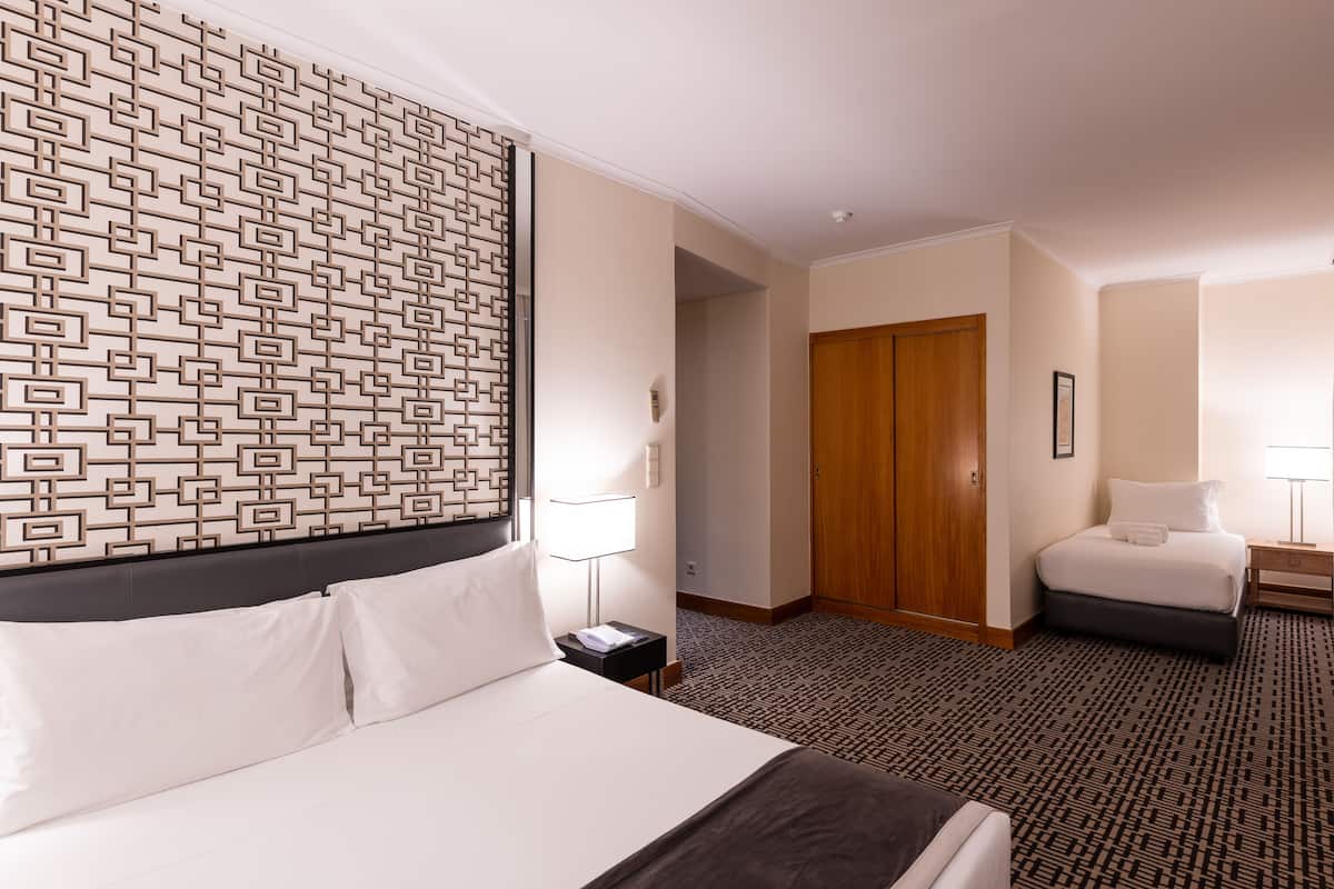 A hotel room with a large bed, a smaller bed, a patterned headboard, a wooden door, and a carpeted floor.