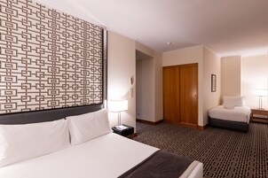 Ett hotellrum med en stor säng, en mindre säng, en sänggavel med mönster, en dörr av trä och ett golv täckt av matta.