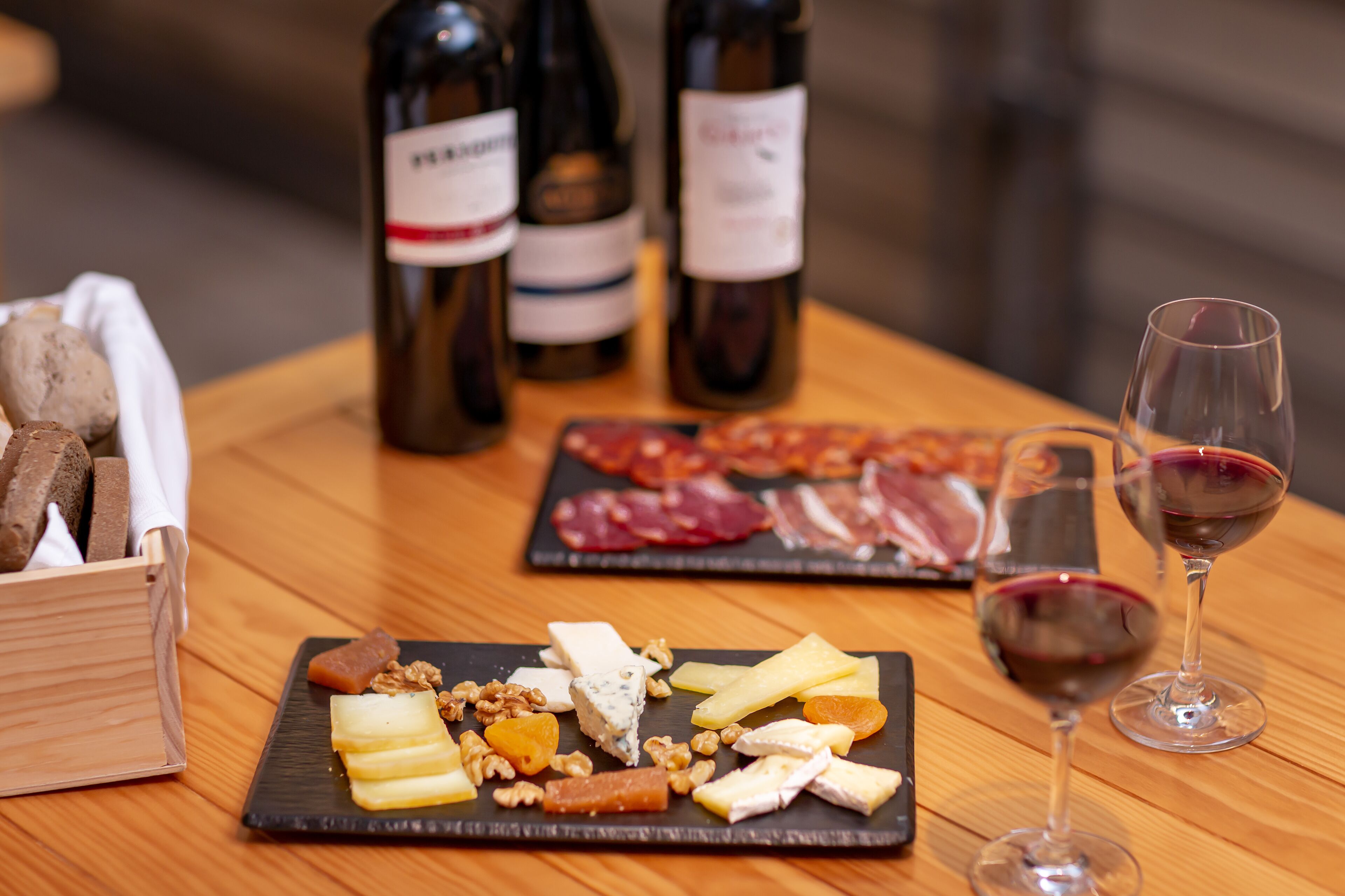 Uma mesa de madeira com tábua de queijos, copos de vinho e garrafas.