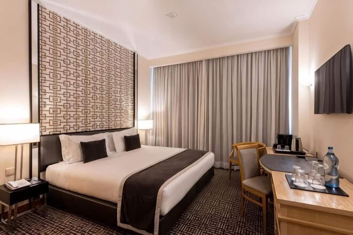 A hotel room with a large bed, a desk with a chair, a television, and a window with curtains.